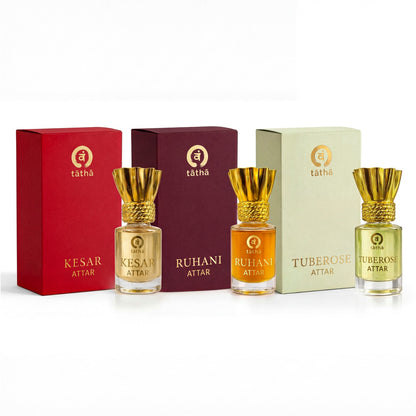 Three attar bottles with gold caps and different colored liquids, each in front of a matching colored box, on a white background.